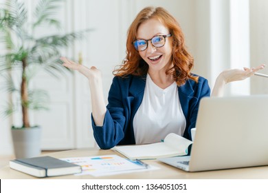 Image Of Cheerful Redhead Businesswoman Watches Webinar Or Tutorial Video, Uses Free Internet Connection, Raises Palms With Hesitation, Sits At Desktop With Papers. Female Owner Of Small Business