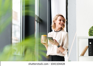 Image Of Cheerful Young Business Woman In Office Looking Aside Chatting By Mobile Phone Drinking Coffee.