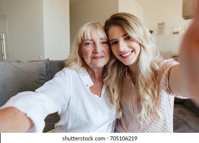 Image Of Cheerful Young Lady Sitting On Sofa At Home With Her Grandmother Hugging. Looking Camera Make Selfie.