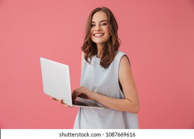 Image of cheerful young woman standing isolated over pink background using laptop computer. Looking camera.
