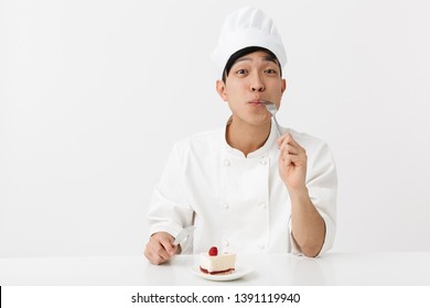 Image Of Cheery Asian Chief Man In White Cook Uniform Smiling At Camera While Eating Tasty Cheesecake Isolated Over White Background