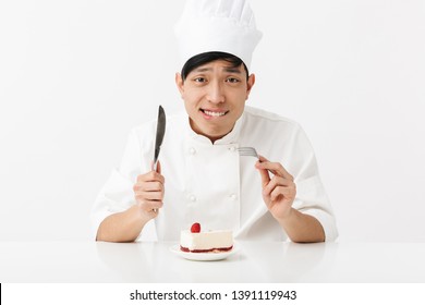 Image Of Cheery Asian Chief Man In White Cook Uniform Smiling At Camera While Eating Tasty Cheesecake Isolated Over White Background