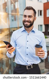 Image Of A Cheery Young Business Man Standing Near Business Center Holding Cup Of Coffee Using Mobile Phone.