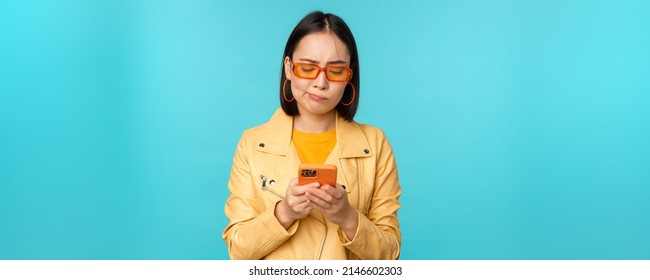 Image Of Chinese Young Woman Looks Disappointed At Her Smartphone, Looking At Mobile Phone With Regret, Standing Over Blue Background