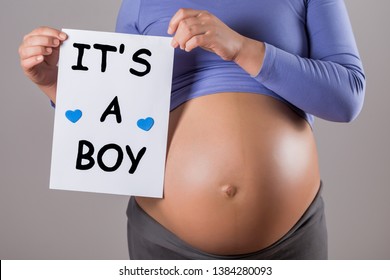 Image of close up stomach of pregnant woman holding paper with text it's a boy on gray background.