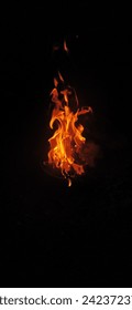 The image is a close-up of fire flames on a black background. The flames are bright orange and yellow, with some wisps of smoke rising from them. The flames are slightly out of focus, giving the image