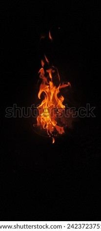 The image is a close-up of fire flames on a black background. The flames are bright orange and yellow, with some wisps of smoke rising from them. The flames are slightly out of focus, giving the image