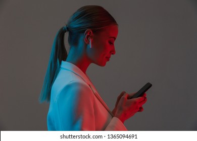 Image Closeup Of Gorgeous Woman 20s Wearing Earpods Holding And Using Mobile Phone While Standing Under Neon Lights Isolated Over Gray Background