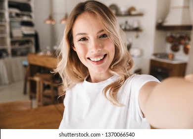 Image closeup of pretty blond woman 20s wearing white t-shirt smiling while looking at camera and taking selfie photo in living room