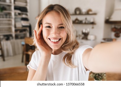 Image closeup of stylish blond woman 20s wearing white t-shirt smiling while looking at camera and taking selfie photo in living room