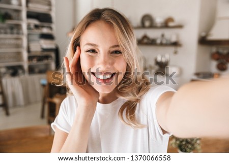Image closeup of stylish blond woman 20s wearing white t-shirt smiling while looking at camera and taking selfie photo in living room