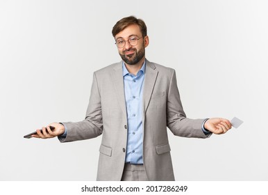 Image Of Clueless Smiling Businessman In Glasses And Gray Suit, Spread Hands Sideways And Shrugging Confused, Holding Mobile Phone And Credit Card