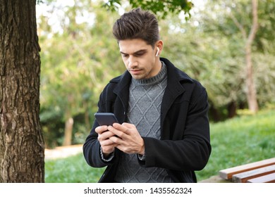 Image of a concentrated handsome young man in casual clothing walking outdoors in green park using mobile phone listening music with earphones.