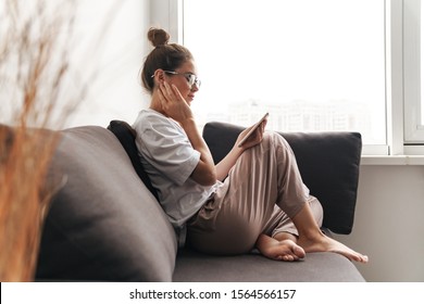 Image of concentrated nice woman in eyeglasses using cellphone while sitting on sofa at living room