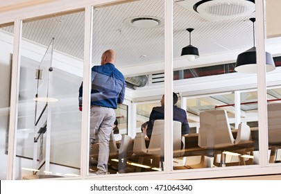 Image Of A Conference Room In A Modern Office Space Taken From Outside While Businessmen And Businesswoman Conduct Their Board Room Meeting Inside.