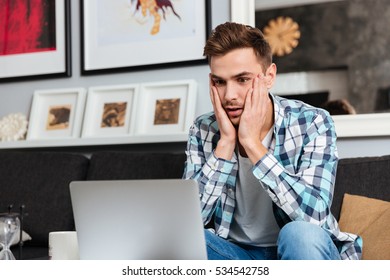 Image Of Confused Bristle Man Dressed In Shirt In A Cage Print Sitting On Sofa In Home And Using Laptop Computer. Looking At Laptop While Holding Face With Hands.