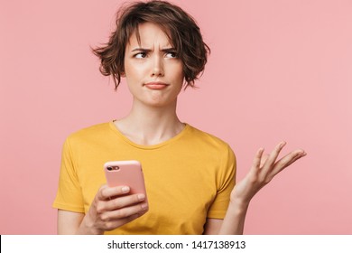Image of a confused young beautiful woman posing isolated over pink wall background using mobile phone.