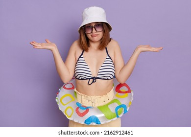 Image of confused young dark haired woman wearing swimsuit and panama posing with rubber ring isolated over purple background, standing shrugging shoulders, being not sure what tour to choose.