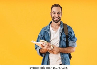Image of content student guy in denim clothes smiling while holding exercise books isolated over yellow background