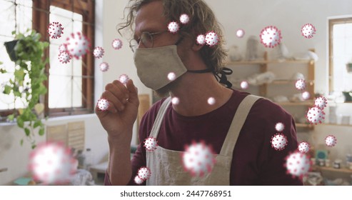 Image of coronavirus cells floating over caucasian man in face mask coughing at pottery workshop. creative business and hobbies during covid 19 pandemic concept digitally generated image. - Powered by Shutterstock