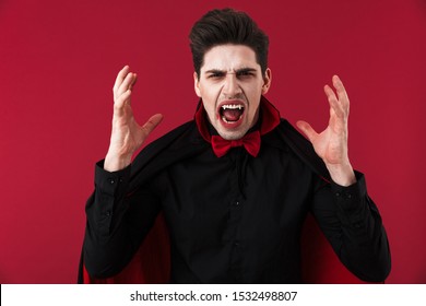 Image Of Crazy Vampire Man With Blood And Fangs In Black Halloween Costume Screaming Isolated Over Red Wall