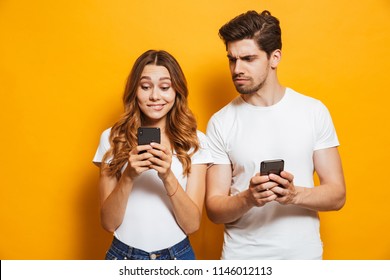 Image of curious man frowning and peeking at smartphone of his girlfriend isolated over yellow background
