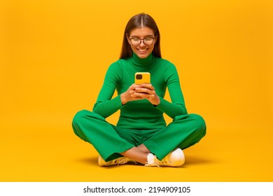 Image Of Cute Attractive Student Girl In Green Clothes Sitting On Floor With Crossed Legs On Yellow Background Using Smartphone Watching Live Podcast, Chatting With Friends