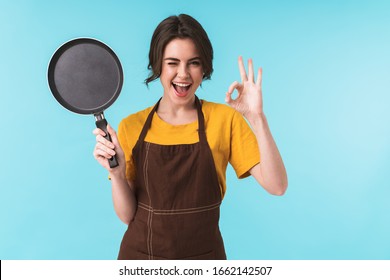 Image of cute happy young woman chef holding frying pan isolated over blue wall background showing okay gesture.