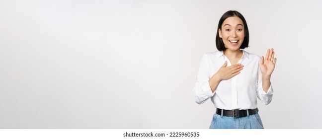 Image of cute young female office worker, asian girl student raising hand up and put palm on chest, name herself, introduce, making promise, standing over white background. - Shutterstock ID 2225960535