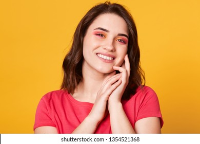 Image of cute young lady standing isolated over yellow background. Looking directly at camera, keeps hands near cheek, has pleasent expression, glad to be photographed. People and lifestyle concept. - Shutterstock ID 1354521368