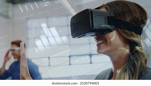 Image of data, charts and digital interface over smiling woman wearing VR headset using virtual screen, with colleague in the background. Global digital network concept digital composite.