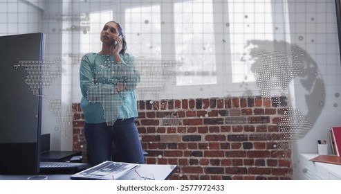 Image of data processing, world map over biracial businesswoman talking on smartphone in office. Global business and digital interface concept digitally generated image.