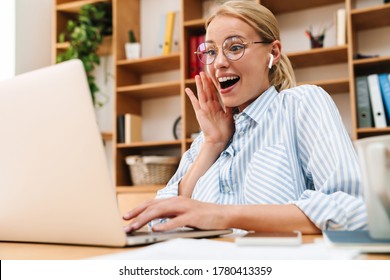 Image of delighted woman using earphones and expressing surprise while working with laptop at table in office