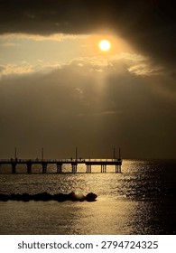 The image depicts a dramatic sunset over the sea, with the sun partially obscured by clouds, casting a golden glow on the water. A pier extends into the sea, silhouetted against the light, with a few people visible. The sky features a mix of dark and illuminated clouds, creating a striking contrast. The sea reflects the sunlight, enhancing the scene's warmth and depth.