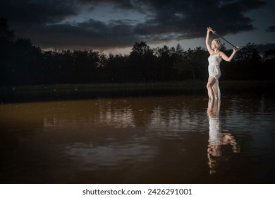 The image depicts an ethereal scene with a young woman in a light, flowy dress standing in a lake at twilight. She is holding a lit sparkler above her head, creating a dynamic contrast with the dusky - Powered by Shutterstock