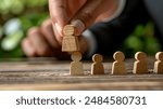 The image depicts a person placing a wooden figure among a row of similar wooden figures, symbolizing talent management, hiring, and human resources strategies.
