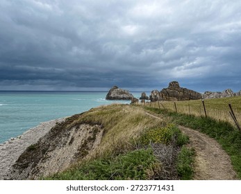 The image depicts a section of the Costa Quebrada, along the Atlantic coastline near Santander, in northern Spain. A narrow dirt path runs along the edge of the cliff, bordered by a simple wooden and metal fence that guides the way without breaking the continuity of the landscape.

To the left, the cliff slopes down toward the turquoise sea, seemingly calm yet marked by subtle movement beneath the surface. Ahead, large isolated rock formations rise from the water, shaped by erosion and time, their layered surfaces revealing the geological history of this stretch of coast. The rocks stand against the horizon as solid, silent presences.

Above, the sky is filled with dense, layered clouds, darker near the horizon, creating a strong contrast with the brightness of the sea. The light is soft and diffused, with no harsh shadows, contributing to a suspended, almost contemplative atmosphere. The scene conveys a sense of walking and anticipation, characteristic of Atlantic landscapes, where time appears to slow down and nature remains the dominant force.