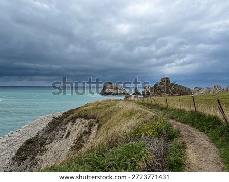 The image depicts a section of the Costa Quebrada, along the Atlantic coastline near Santander, in northern Spain. A narrow dirt path runs along the edge of the cliff, bordered by a simple wooden and metal fence that guides the way without breaking the continuity of the landscape.

To the left, the cliff slopes down toward the turquoise sea, seemingly calm yet marked by subtle movement beneath the surface. Ahead, large isolated rock formations rise from the water, shaped by erosion and time, their layered surfaces revealing the geological history of this stretch of coast. The rocks stand against the horizon as solid, silent presences.

Above, the sky is filled with dense, layered clouds, darker near the horizon, creating a strong contrast with the brightness of the sea. The light is soft and diffused, with no harsh shadows, contributing to a suspended, almost contemplative atmosphere. The scene conveys a sense of walking and anticipation, characteristic of Atlantic landscapes, where time appears to slow down and nature remains the dominant force.