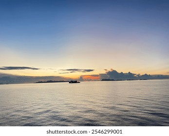 The image depicts a serene seascape at dawn. The sun is rising, casting a warm glow on the horizon and painting the sky in hues of orange, pink, and blue. The calm ocean reflects the sky's colors, cre - Powered by Shutterstock