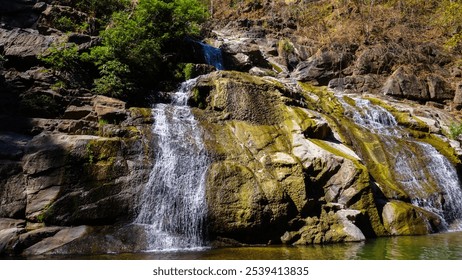 The image depicts a serene waterfall cascading over rocky terrain, surrounded by lush greenery. The water flows gently into a calm pool below. - Powered by Shutterstock