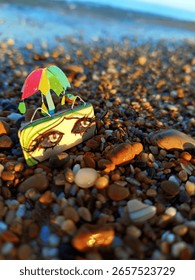 The image depicts a small, decorative object resembling a stylized suitcase with painted eyes and a small, colorful umbrella, resting on a pebble beach. The background features the ocean and the horiz