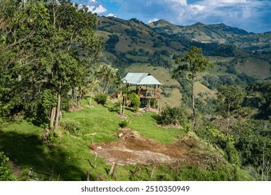 The image depicts a small, isolated house situated on a hillside in a rural, mountainous area. The house, which has a metal roof, is surrounded by lush greenery, including tall trees and dense vegetat - Powered by Shutterstock