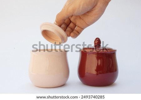 The image depicts two ceramic containers or cups. One container features a beige exterior with a lid, while the other is held by a human hand, revealing its maroon exterior and glossy finish. 