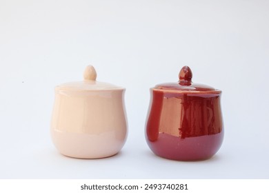 The image depicts two ceramic pots with lids. One pot is glazed in a light cream color, while the other features a glossy maroon finish. Both pots have a round body, a flat base, and an ornamental kno