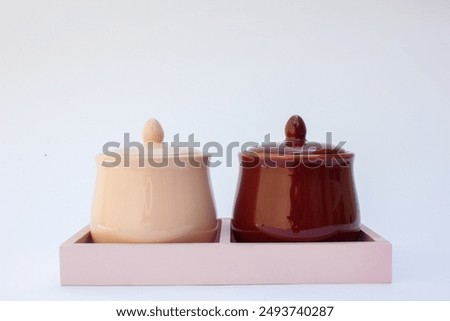 The image depicts two ceramic pots with lids. One pot is glazed in a light cream color, while the other features a glossy maroon finish. Both pots have a round body, a flat base, and an ornamental kno