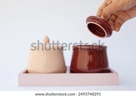 The image depicts two ceramic pots with lids. One pot is glazed in a light cream color, while the other features a glossy maroon finish. Both pots have a round body, a flat base, and an ornamental kno