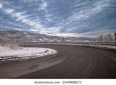 The image depicts a winding mountain road on a cold winter day, with snow lining the asphalt and guardrails. The scene is set against a backdrop of gently rolling, snow-covered hills and a dramatic sk - Powered by Shutterstock