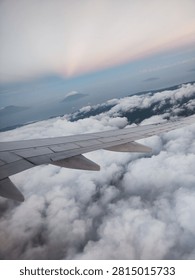 Image Description
 
This is a view captured from an airplane window while flying high above the clouds. ✈️
 
The metallic grey wing of the aircraft stretches across the lower half of the frame, extending out toward the horizon. Below lies a vast, thick sea of fluffy white clouds, billowing and stretching endlessly like soft cotton. Peeking through gaps in the cloud cover in the distance are the dark, hazy silhouettes of mountain peaks and islands rising above the horizon.
 
The sky above shows a gentle gradient: pale misty blue shifts smoothly into faint pastel pink and soft lavender near the top, suggesting the soft light of early dawn or late twilight. The whole scene feels calm, airy, and majestic, giving a wonderful sense of floating high above the world below. ☁️🏔️