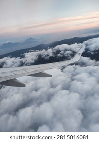 Image Description
 
This is a view captured from an airplane window while flying high above the clouds. ✈️
 
The metallic grey wing of the aircraft stretches across the lower half of the frame, extending out toward the horizon. Below lies a vast, thick sea of fluffy white clouds, billowing and stretching endlessly like soft cotton. Peeking through gaps in the cloud cover in the distance are the dark, hazy silhouettes of mountain peaks and islands rising above the horizon.
 
The sky above shows a gentle gradient: pale misty blue shifts smoothly into faint pastel pink and soft lavender near the top, suggesting the soft light of early dawn or late twilight. The whole scene feels calm, airy, and majestic, giving a wonderful sense of floating high above the world below. ☁️🏔️