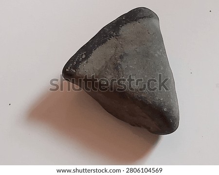 The image displays a solid, triangular or trapezoidal object made of dark stone. Its surface appears smooth and polished, which is a characteristic of technology from the Neolithic period (New Stone Age). This object shares features with a celt or a ground-stone axe, a tool that was ground and sharpened on one end to be used for daily activities such as wood-working or farming. These types of artifacts were typically mounted onto wooden handles to serve as effective chopping tools.